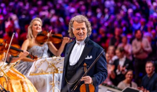 A man with long curly hair stands on a stage holding a violin Behind him is a large crowd clapping