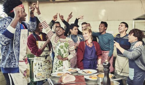 A middleaged woman in an apron is surrounded by youths celebrating in a kitchen