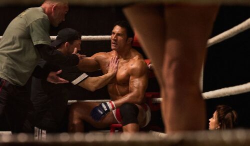 A muscular man sits in the corner of a boxing ring being held back by his coach In the foreground is a pair of legs