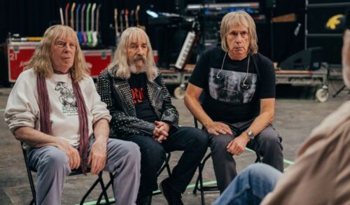Three old men wearing leather jackets and band tshirts sit on chairs in the backstage of a music venue