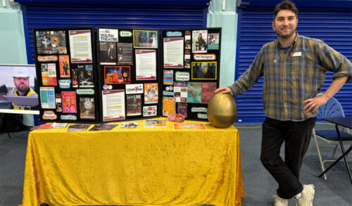 A man in a plaid shirt standing next to a stall with information boards and giant golden egg