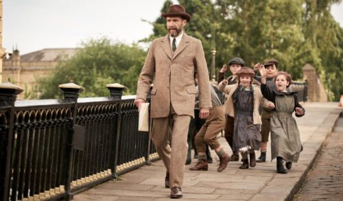 A man in a brown suit and hat walks down the street as a group of small children shout behind him