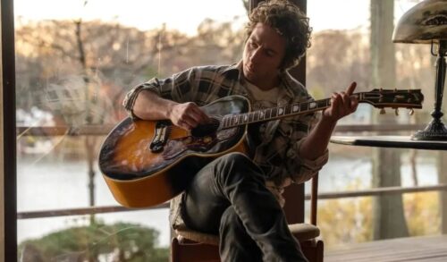 A man sits on a chair whilst playing an acoustic guitar