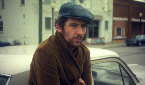 A man in a brown coat and grey flat cap leans next to a car