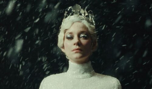A woman with white hair a crown and a white jumper walks through a snow storm