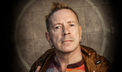 A man with short spiky blonde hair earrings and a brown jacket looks at the camera