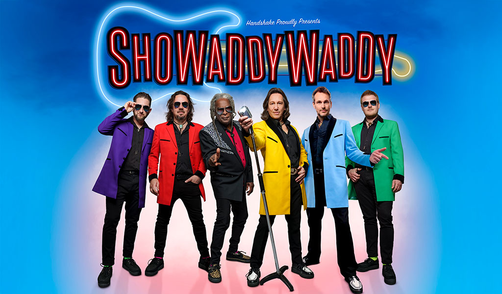 A group of men in colourful suits posing with a microphone. The text about them reads Showaddywaddy’ with a neon guitar behind them.