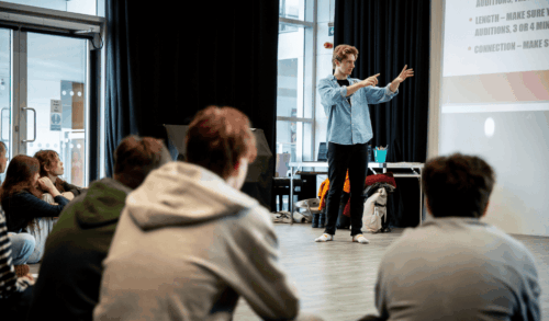 A man in a blue shirt holding arms up standing in front of young people sat on the floor
