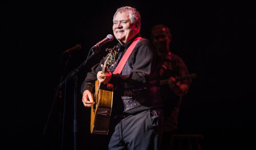 An older gentleman playing a guitar and singing into a microphone on stage