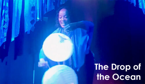 A photo of a woman performing on stage holding two reflective circles There are blue streamers hanging behind her The text in the bottom right reads The Drop of the Ocean