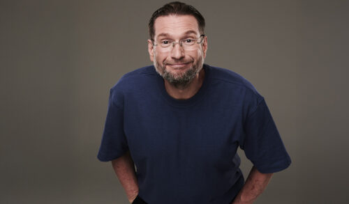 The comedian Gary Delaney  wearing a blue tshirt and glasses leas forward and smiles at the camera with his hands in his pockets
