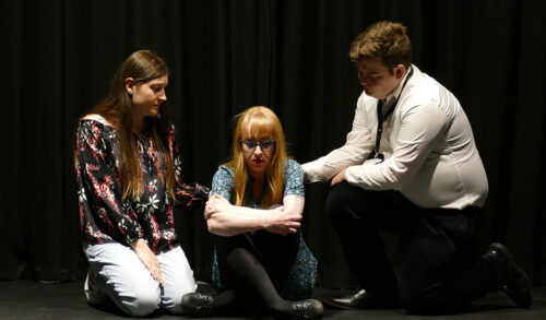 A photo of a blonde woman sat on the floor hugging her knees with a man and a woman kneeling next to her with their hands on her back