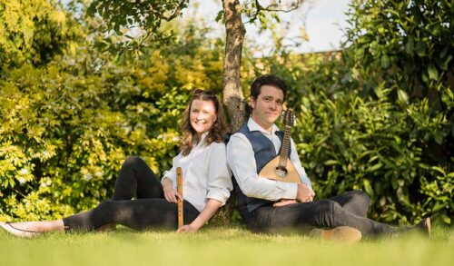 A man and a woman sat with their backs against a tree They are holding a flute and lute