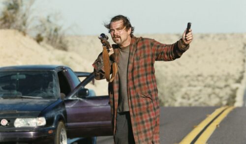 Leonardo Di Caprio in a brown check coat standing next to a car pointing a rifle in one hand and holding a tracking device in the other