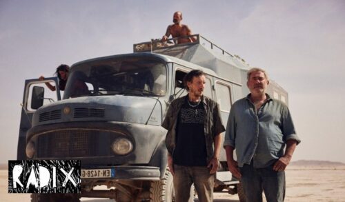 Two men stand in front of a large truck in the middle of a desert Another person is sitting on top of the truck