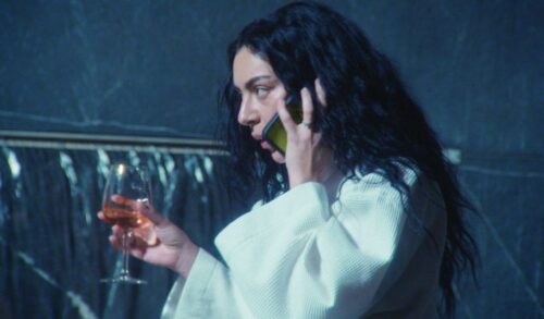 Popstar Charli xcx stands in a white robe talking on the phone whilst she holds a glass of wine