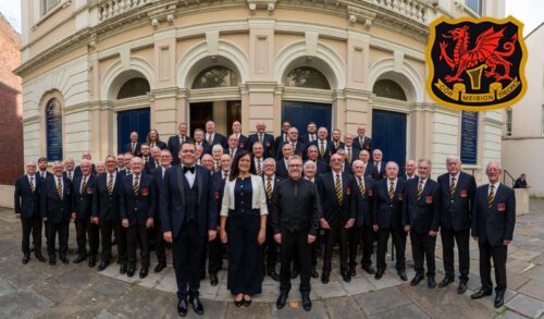 A photo of a group of men and one woman stood outside a building They are all wearing navy blue suits The logo in the top corner has a red dragon and the text underneath reads Cor Meibion Treorci