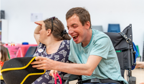 A young man in a wheel chair reaches for something laughing A woman sits next to him looking off into the distance