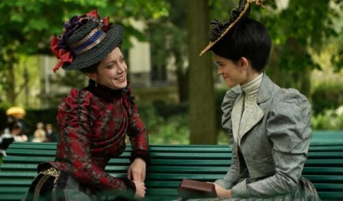 Two women in 19th century dresses and hats sit on a park bench