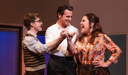 Two men and a woman link hands and smile on stage