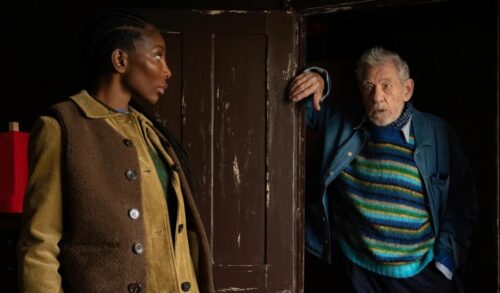 An elderly man in a blue stripy jumper leans on a doorway across from a woman in a brown coat