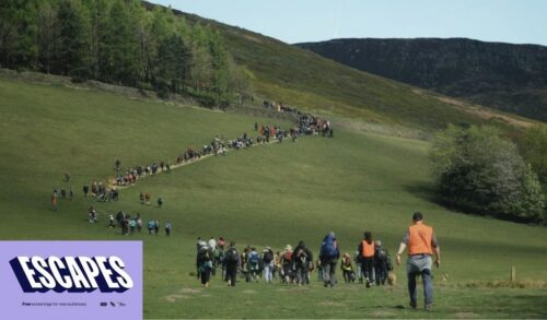 A large field on a hill with a line of protestors walking up it In the bottom left is a logo that reads Escapes